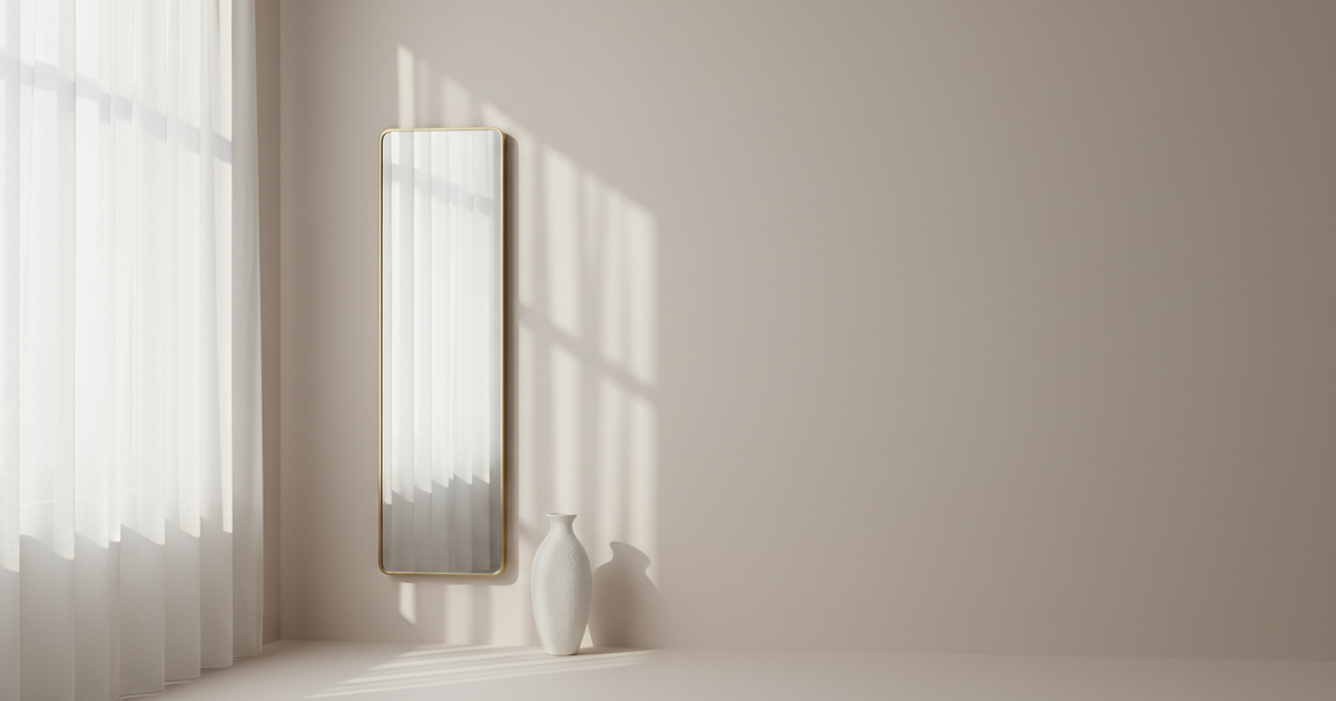 Halbtransparente Gardine vor einem Fenster diffundiert Tageslicht, ein schlanker Wandspiegel und eine weiße Keramikvase.