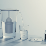 Minimalistisches Stillleben mit Wasserfilterkanne, die in ein klares Trinkglas gießt, daneben Petrischale und Aktivkohlegranulat als Funktionshinweis