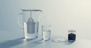 Minimalistisches Stillleben mit Wasserfilterkanne, die in ein klares Trinkglas gießt, daneben Petrischale und Aktivkohlegranulat als Funktionshinweis