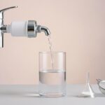 Wasserhahn mit Aktivkohlefilter füllt eine klare Glaskaraffe, daneben Labortrichter und feines Sieb, viel Negativraum.