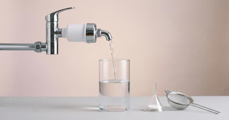 Wasserhahn mit Aktivkohlefilter füllt eine klare Glaskaraffe, daneben Labortrichter und feines Sieb, viel Negativraum.