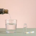 Minimalistisches Studio Stillleben mit altem Metall Wasserrohrabschnitt, aus dem ein feiner Wasserstrahl in ein klares Trinkglas fließt, daneben Probefläschchen und Teststreifen.