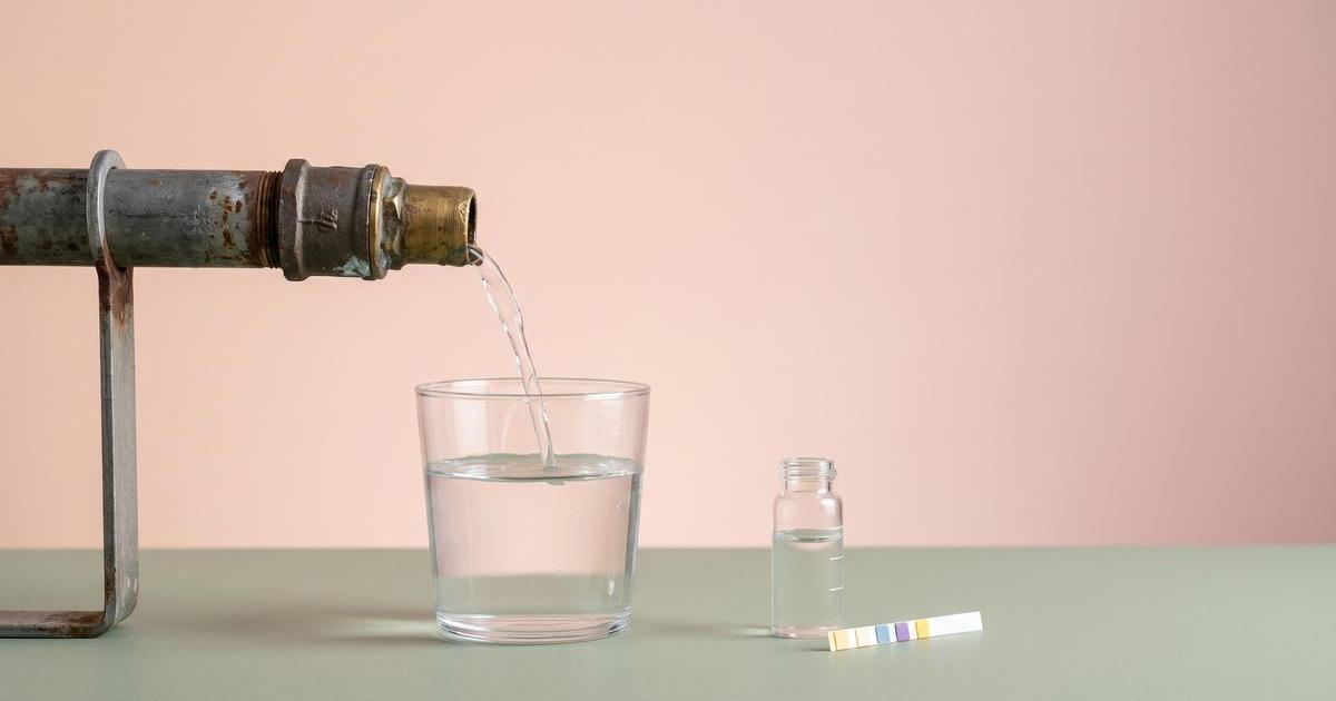 Minimalistisches Studio Stillleben mit altem Metall Wasserrohrabschnitt, aus dem ein feiner Wasserstrahl in ein klares Trinkglas fließt, daneben Probefläschchen und Teststreifen.