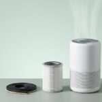 Minimalistisches Studio-Stillleben mit laufendem Luftreiniger und sichtbarem HEPA-Filter, daneben ein Ersatzfilter und eine aufgerollte Fensterdichtung.