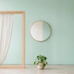Minimalistisches Stillleben mit Fensterrahmen und halbtransparentem Vorhang, ein Lichtkegel fällt auf pastelligen Hintergrund, Spiegel und Zimmerpflanze als Kontext.
