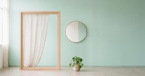 Minimalistisches Stillleben mit Fensterrahmen und halbtransparentem Vorhang, ein Lichtkegel fällt auf pastelligen Hintergrund, Spiegel und Zimmerpflanze als Kontext.