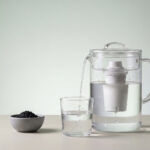 Minimalistisches Stillleben mit Tischwasserfilter, der Leitungswasser in ein Glas filtert, daneben Aktivkohlegranulat als Hinweis auf PFAS Reduktion.