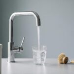 Küchenarmatur füllt ein Trinkglas, daneben Reinigungsbürste und Filterkartusche, minimalistisches Studio Stillleben mit viel Negativraum