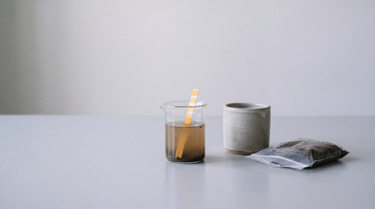 Studio-Stillleben eines pH-Bodentest-Sets mit Erd-Wasser-Becherglas, Probenbeutel und hellgrauem Keramiktopf, symbolisiert sichere Bodenanalyse.